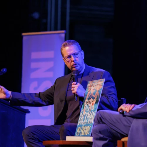Presenter on stage speaking to audience under spotlight.