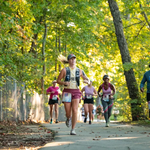 A group of runners heading through the woods with race numbers and pretty, well-lit trees in the background.