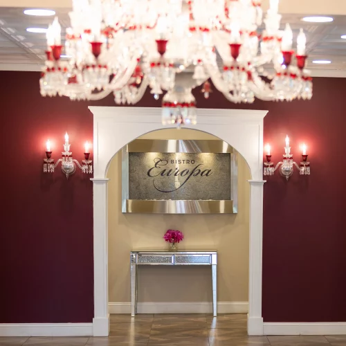 Interior view of a refined entryway featuring rich maroon accent walls, elegant wall sconces, a crystal chandelier, and a decorative console table beneath an arched doorway.