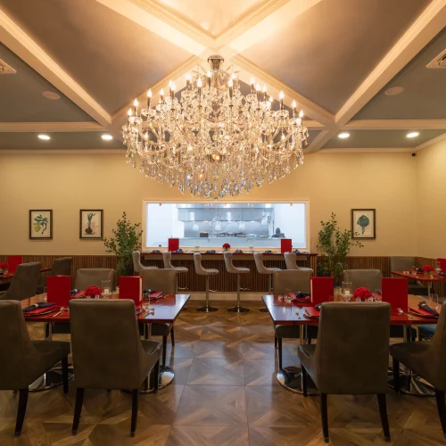 Wide-angle view of a modern dining room featuring a large crystal chandelier, neutral walls, elegant table settings, and an open kitchen window framed by warm wood accents and wall art.