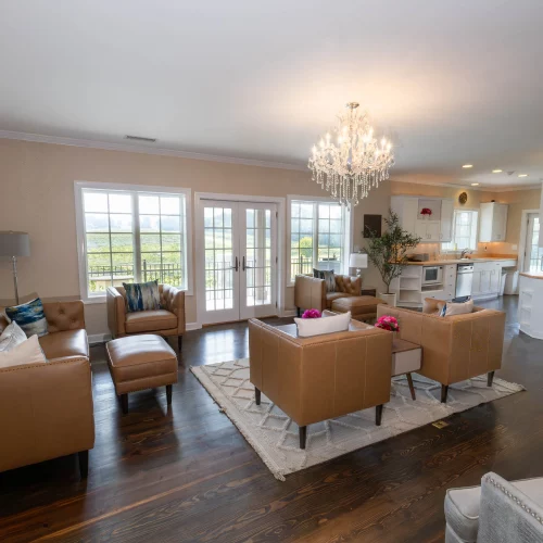 Bright open-concept living room with tan leather furniture, a crystal chandelier, hardwood floors, and large windows leading to a scenic outdoor view.