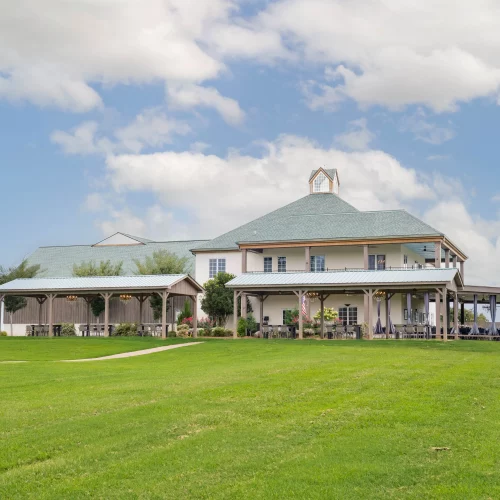 Exterior photograph of a large countryside estate with wraparound patios, manicured lawn, and a bright blue sky with scattered clouds.